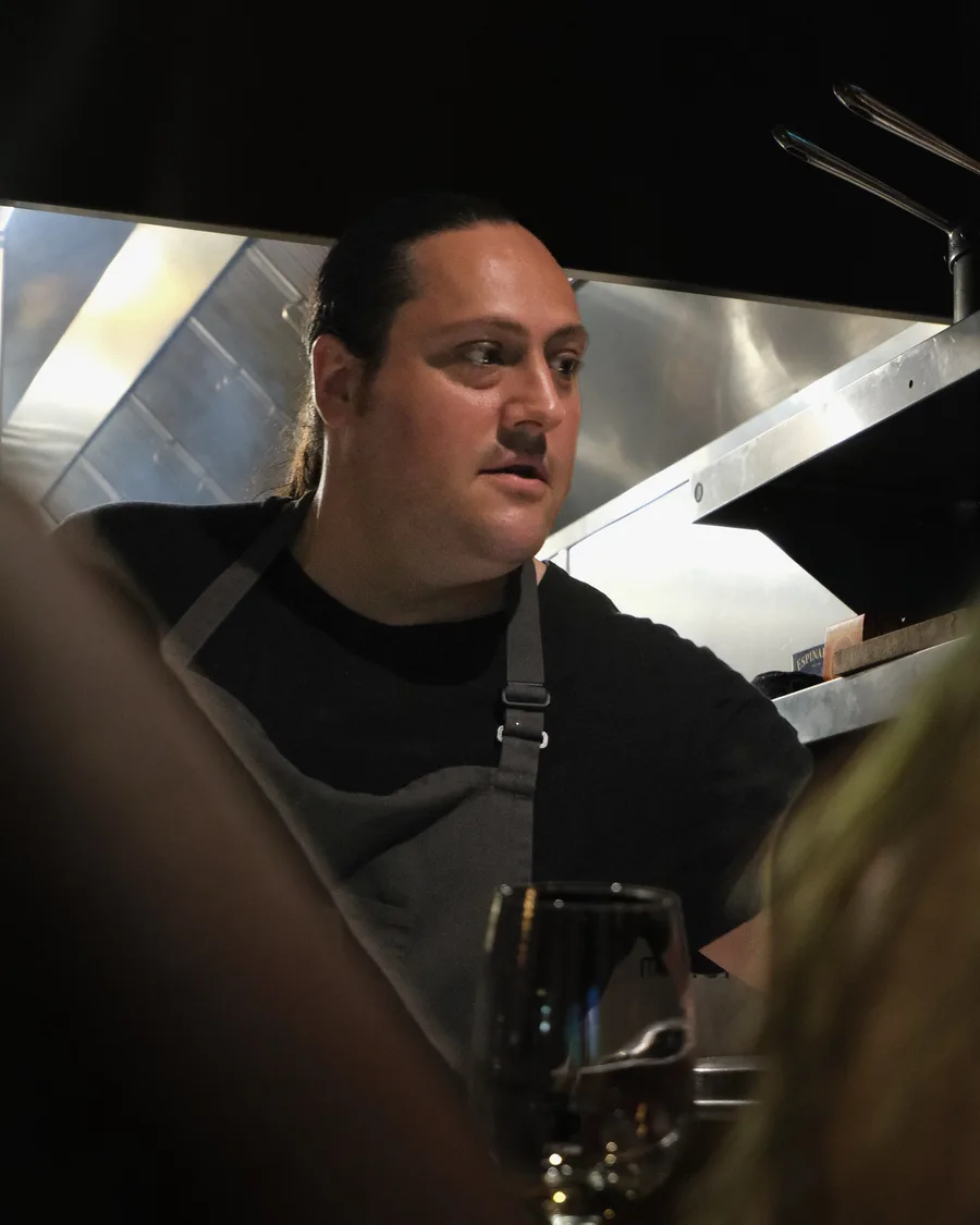 Chef Kyle Romanowski in his apron, talking with guests at the pass